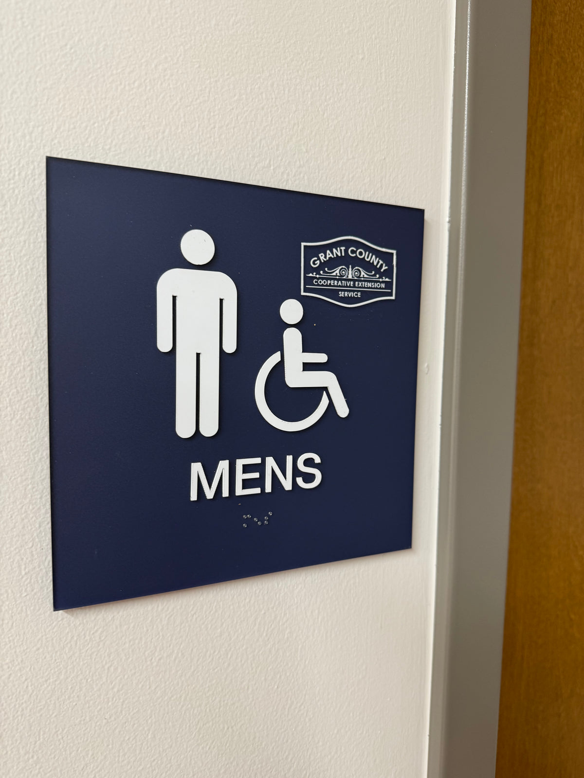 Blue ADA men's restroom sign with raised white lettering, accessibility symbols and braille for Grant County Cooperative Extension.
