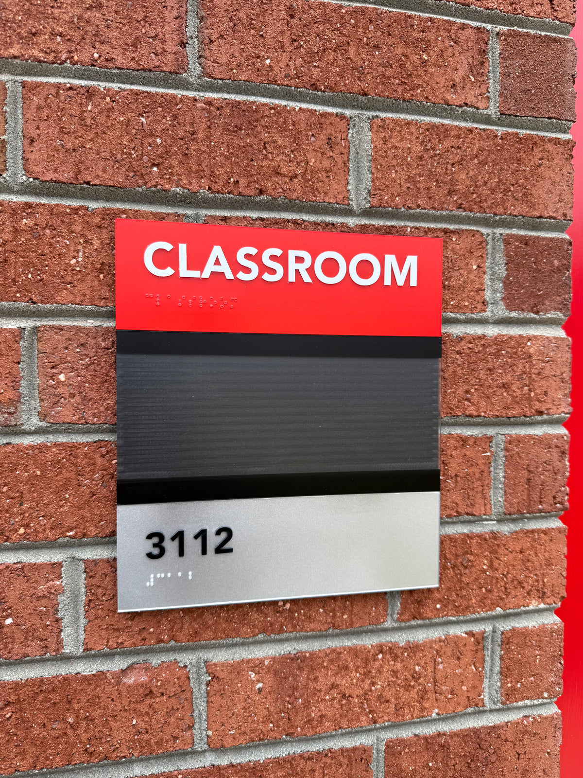 Red, black and silver classroom identification sign with raised lettering, room number, braille and changeable message area mounted on a brick wall.