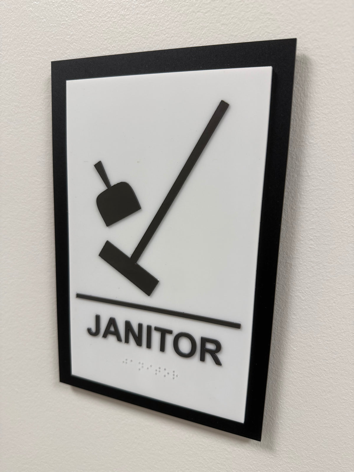 Black and white janitor room sign with raised pictogram, tactile lettering and braille.