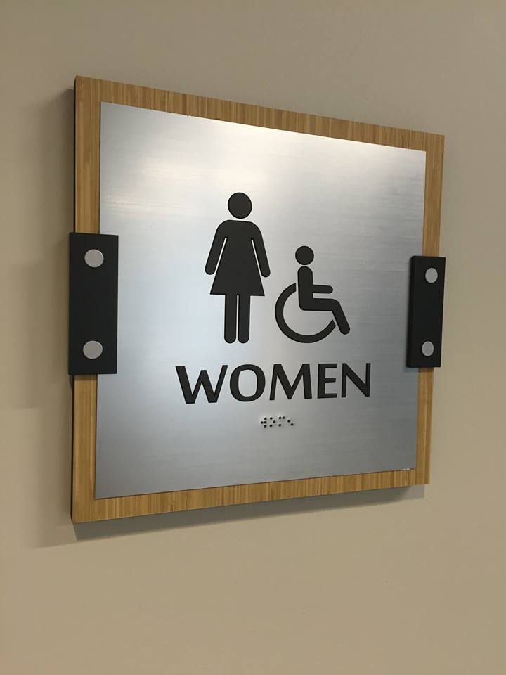 Custom women's ADA restroom sign with brushed metal face, wood-look backer, raised pictograms, tactile lettering, and braille.